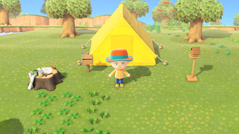 A villager in an orange wide brim hat with a blue rim, orange and yellow flannel, blue jeans, and yellow loafers standing in front of yellow tent outside with grass, weeds, and trees in the background. The crafting station is to the left of the villager and the birdhouse is to the left of the villager. There are plants in front of the villager.